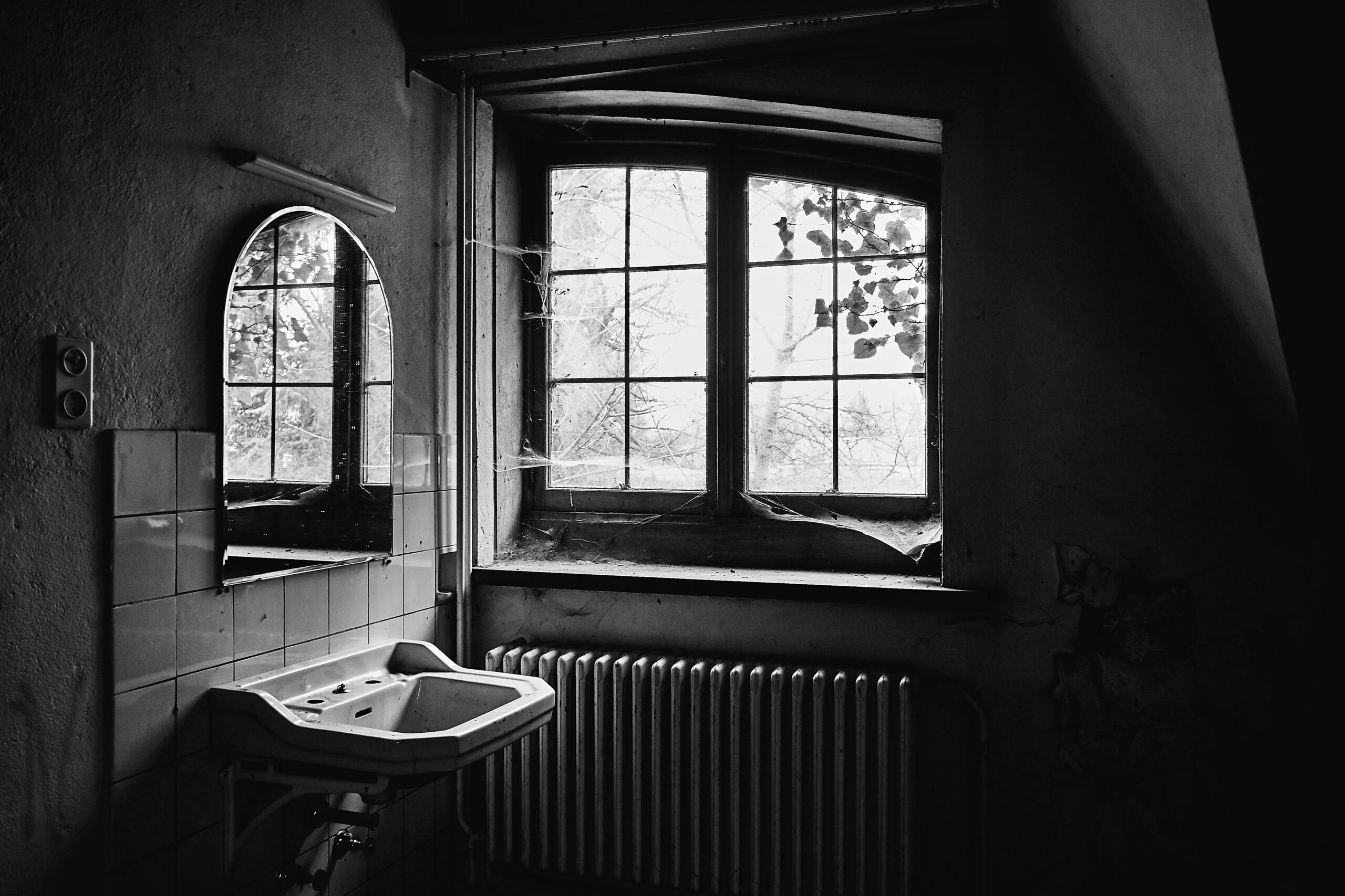 A grayscale shot of an abandoned room with a sink and a mirror and spiderwebs all over the window